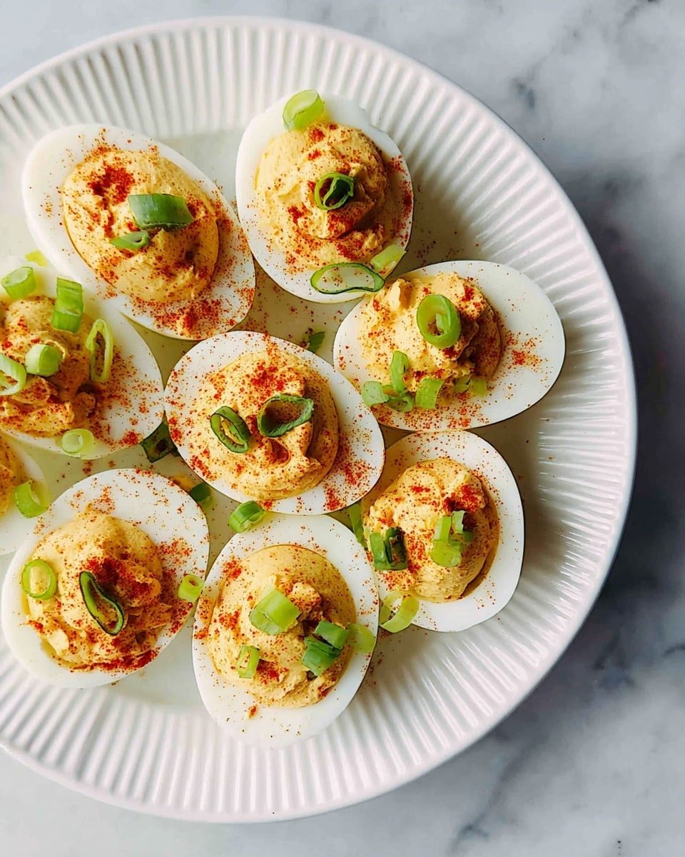 A white, round plate with ridged edges holds twelve deviled eggs arranged closely together. Each egg half has a smooth white outer layer of egg white, filled with a creamy, light yellow yolk mixture that has a slightly rough texture on top. The yolk filling is sprinkled with reddish-brown paprika and garnished with thin slices of bright green scallions, adding color contrast. The plate sits on a white marbled surface. photo taken with an iphone --ar 4:5 --v 7