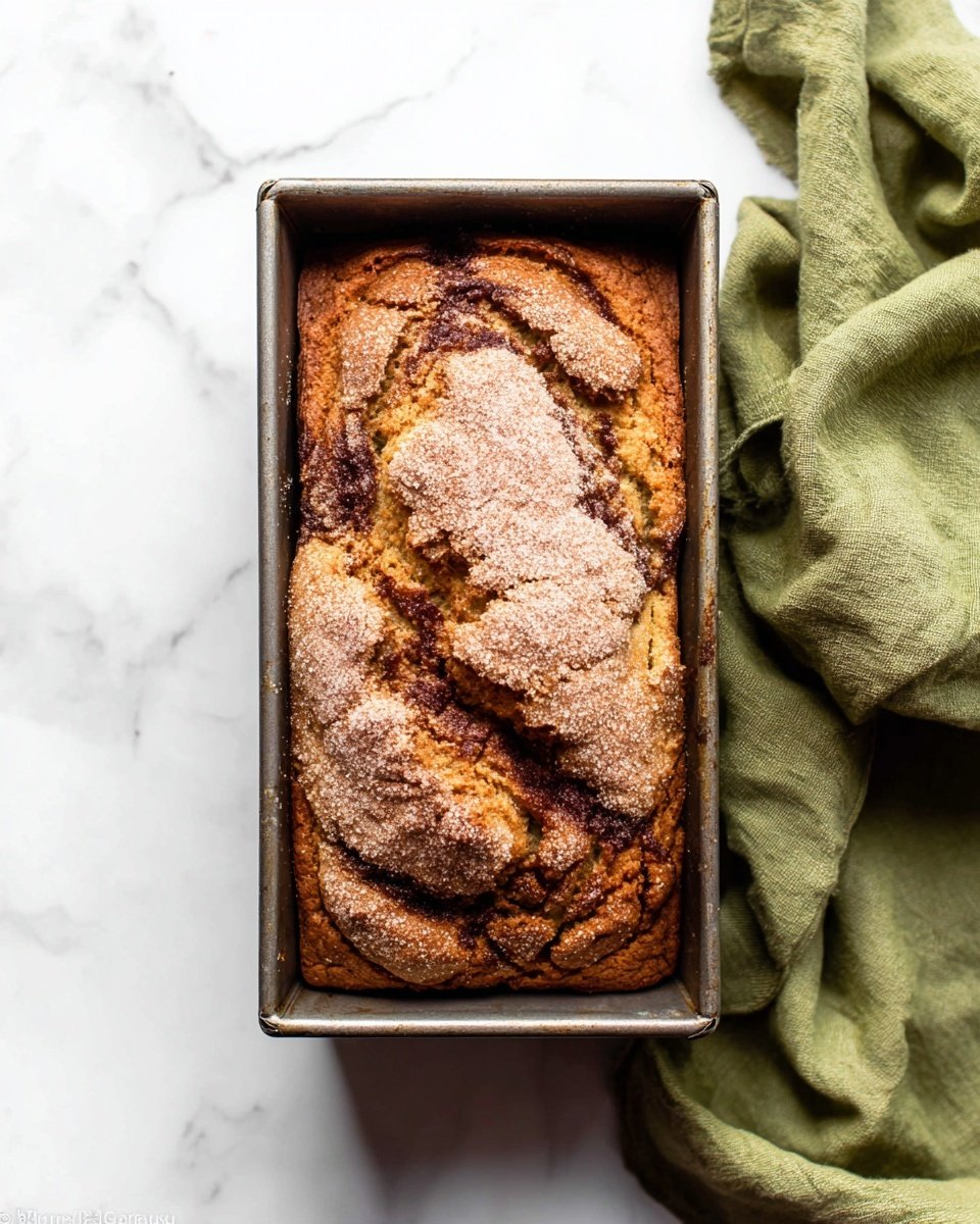 A loaf of baked bread in a metal rectangular pan sits on a white marbled surface. The bread has two main layers, a golden-brown top with a slightly cracked, rough texture laid with light sugar or cinnamon dust, and a darker brown, almost cinnamon swirl layer visible through the cracks. The top has a slightly uneven, rustic crust with some rises and deeper cracks, showing its soft inside. A soft green cloth with folds is placed to the right of the pan. photo taken with an iphone --ar 4:5 --v 7