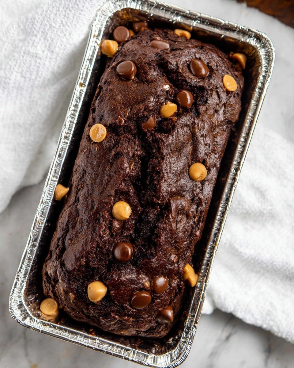 A thick, dark brown chocolate bread with a rough, cracked top sits in a small rectangular silver foil baking pan. Scattered on the surface are smooth, round chocolate chips and light brown peanut butter chips, adding texture and color contrast. The bread has a moist and dense look with visible cracks and uneven grooves running across the top. It rests on a white marbled surface with a white cloth slightly visible nearby. photo taken with an iphone --ar 4:5 --v 7